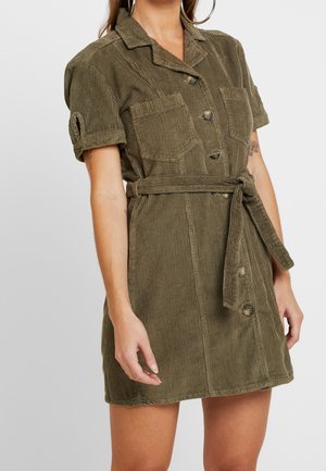 Olivenfarvet corduroy skjortekjole med korte ærmer, to brystlommer, knaplukning foran og en bundet taljebælte for en tætsiddende effekt.