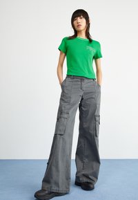 T-shirt verde in cotone con colletto bianco e logo, abbinato a pantaloni cargo grigi a gamba larga con più tasche e dettagli con bottoni.