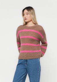 Brązowy sweter z dzianiny z różowymi paskami, okrągły dekolt, luźny krój i teksturowane wykończenie, zestawiony z niebieskimi dżinsami, na jasnym tle.