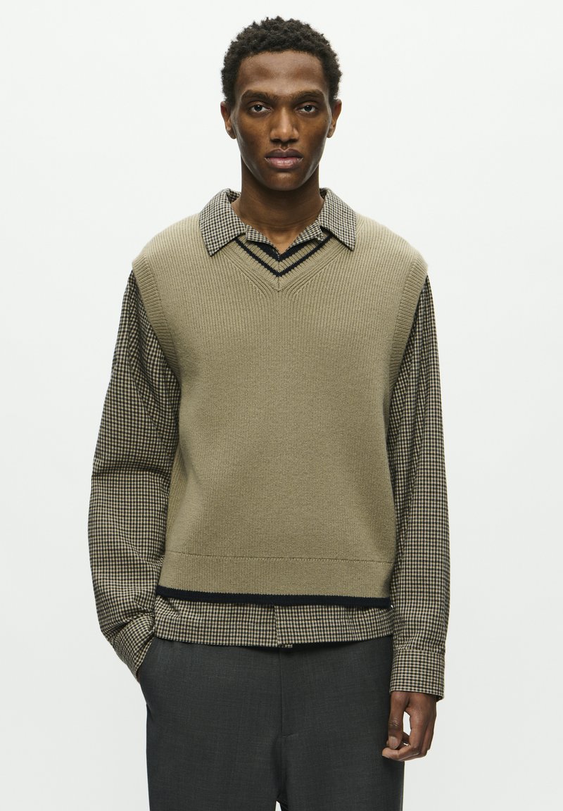 Gilet beige lavorato a maglia con scollo a V sopra una camicia a quadri, caratterizzato da dettagli neri. Il modello indossa pantaloni grigio scuro. Strati e motivi texture.