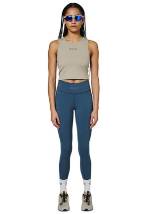 Hellgrünes, cropped Tanktop mit "HALO"-Logo, kombiniert mit hochgeschnittenen marineblauen Leggings. Das Modell trägt außerdem blaue Sonnenbrillen und beigefarbene Sneakers.