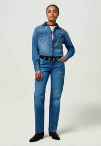 Camisa de denim y jeans en azul medio. La camisa presenta bolsillos puntiagudos y botones de presión. Cinturón negro con hebilla dorada. Botines negros hasta el tobillo.