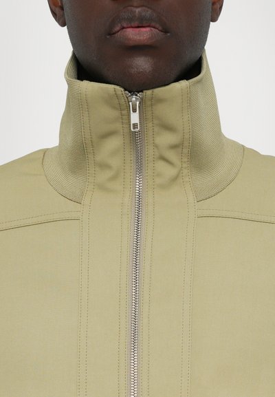 Beige high-collar jacket with a front zipper, textured panel detailing, and visible stitching, made from smooth fabric.