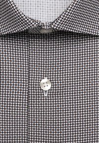 Chemise à col avec un petit motif géométrique en losange noir, blanc et marron, avec un bouton blanc au centre.