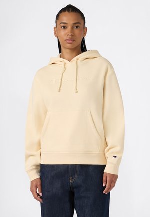 Vrouw draagt een lichtbeige Champion hoodie met een voorkzak en donkerblauwe jeans, staand tegen een effen witte achtergrond.