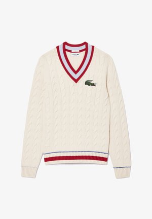 Kermansävyinen palmikkoneulepusero, jossa on punaiset ja siniset raidalliset V-kaula-aukko, hihansuut ja helma sekä vihreä Lacosten krokotiililogo rinnassa.