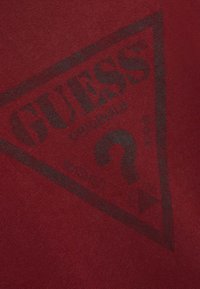 Rode stof met een groot zwart driehoekig Guess-logo en de tekst "GUESS ORIGINALS WASHED" met een gestileerd vraagteken in het midden.