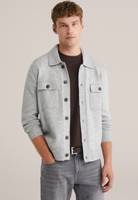 Grå stickad cardigan med knäppning fram, två bröstfickor, ribbade manchetter och krage, bärs över en mörk skjorta.