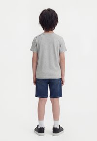 Copil cu păr închis la culoare, privind cu spatele, purtând un tricou gri deschis cu mâneci scurte, pantaloni scurți din denim închis, șosete albe și adidași negri.