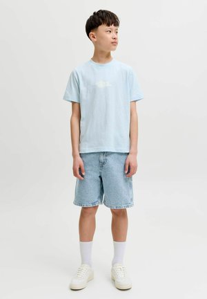Nastolatek ubrany w jasnoniebieski t-shirt z nadrukiem chmur, dżinsowe szorty, białe skarpetki i białe trampki, stojący na jednolitym białym tle.