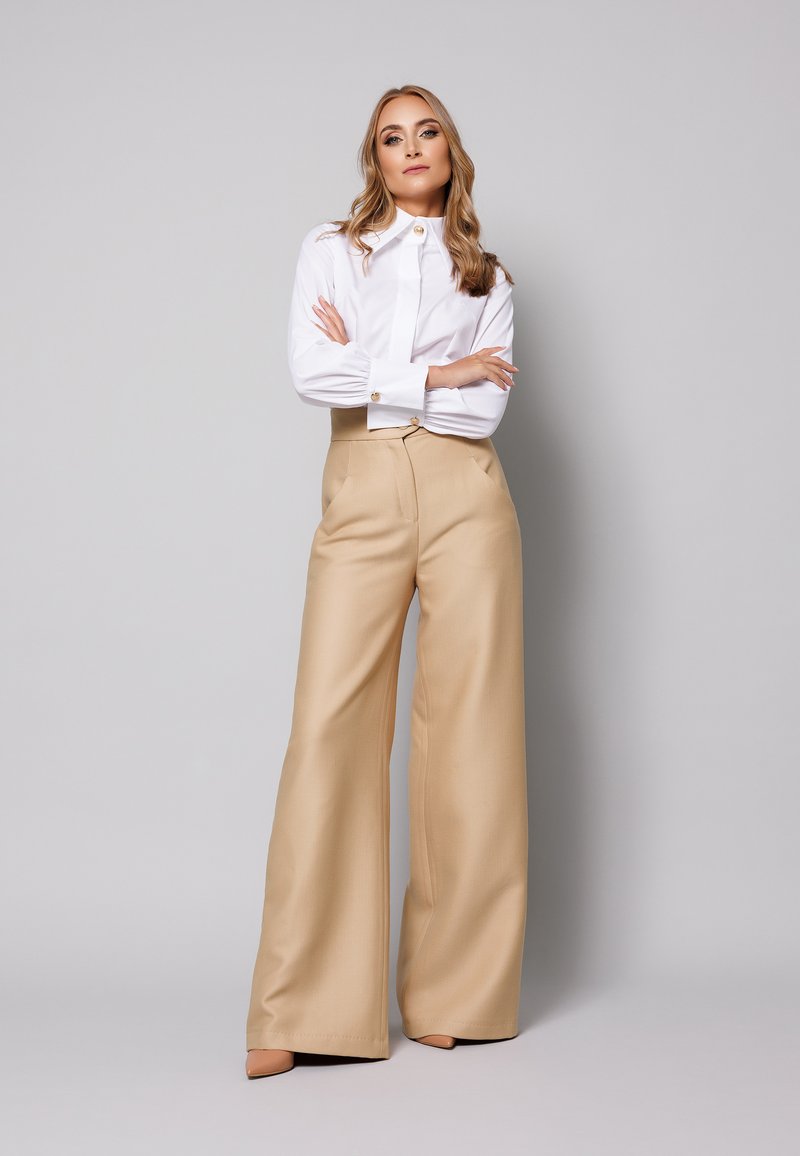 Biała koszula z kołnierzykiem i złotymi guzikami, połączona z beżowymi spodniami o wysokim stanie i szerokich nogawkach, wykonana z gładkiego materiału, bez widocznych wzorów.