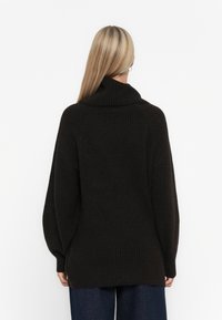 Dunkel oversized Strickpullover mit hohem Kragen, gerippter Textur und langen Ballonärmeln, kombiniert mit locker sitzenden blauen Jeans.