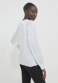 Blusa bianca plissè con maniche lunghe, caratterizzata da un colletto rotondo e polsini elasticizzati, abbinata a pantaloni neri, su uno sfondo chiaro.