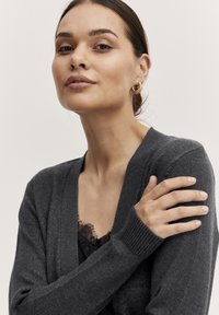 Grå, stickad kofta med V-ringning, ribbade ärmslut och textilstruktur. Buren över en svart spetsblus, med en naturlig, neutral bakgrund.