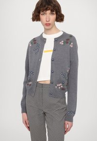 Cardigan en tricot gris avec broderie florale en rose et bleu, col rond et fermeture à boutons. Assorti avec un pantalon à motifs.