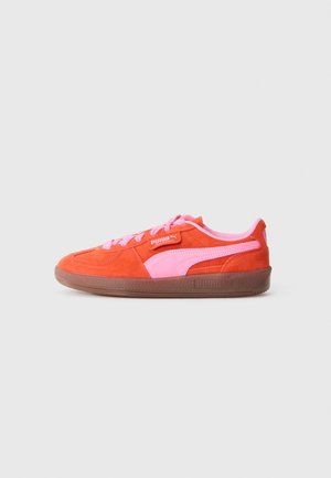 Roter Wildleder-Sneaker mit pinkem Logo-Streifen, pinken Schnürsenkeln und brauner Gummisohle, seitlich auf hellem Hintergrund dargestellt.