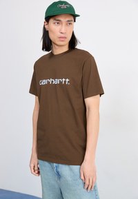 Hnedé bavlnené tričko s bielym logom "carhartt". Skombinované so svetlomodrými džínsami a zelenou čiapkou s vyšitým textom.