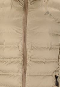 Beige puffjacka med vertikalt quiltade sektioner, en central dragkedja med en brun dragknapp och en liten logotyp i det övre hörnet.