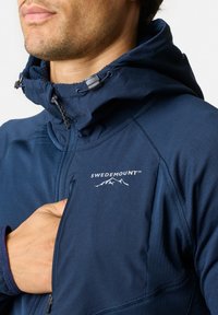 Marineblauwe jas met een ritszak, voorzien van een hoge kraag, verstelbare capuchon en een wit "SWEDEMOUNT" logo met bergdetails.