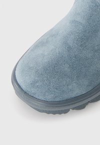Egy kék velúrból készült cipő orrának közeli képe, amelyen egy textúrázott fekete gumitalp látható, fehér háttér előtt.