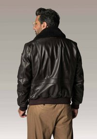 Homme portant une veste en cuir marron foncé avec un col en fourrure noire et un pantalon beige, tourné le dos contre un fond neutre.