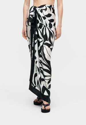 Kobieta nosząca sarong w dużym czarno-białym liściastym wzorze, przewiązany w talii, z sandałami na platformie na jednolitym tle.