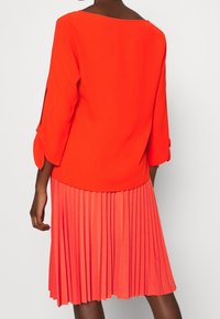 Robe rouge vif avec une jupe plissée, des manches trois-quarts, un col arrondi et une texture lisse. Vue arrière présentant la silhouette fluide.