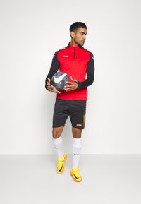 Rode sportjack met ritssluiting en zwarte mouwen, zwarte shorts met oranje accenten, witte sokken en gele Nike-schoenen, terwijl hij een zwarte voetbal vasthoudt.