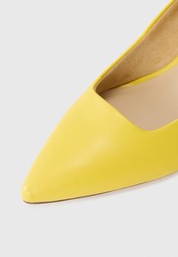 Gros plan sur le bout pointu et la doublure intérieure d'une chaussure plate en cuir jaune sur un fond clair.