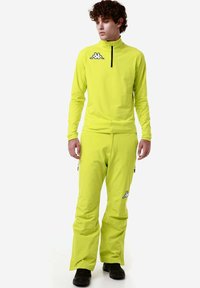 Completo sportivo giallo composto da una maglietta a maniche lunghe con zipper e logo, abbinata a pantaloni coordinati. Il tessuto appare liscio e traspirante.