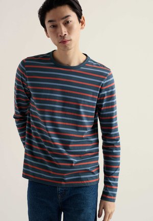 Tričko s dlhým rukávom a okrúhlym výstrihom, ktoré má horizontálne pruhy v modrej, sivej a červenej farbe, vyrobené z mäkkej bavlnenej látky, s voľným strihom.