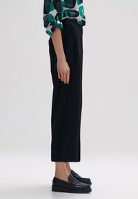 Pantalons noirs ajustés avec une finition texturée, associés à des mocassins noirs élégants. La tenue comprend un haut à motifs verts et blancs.