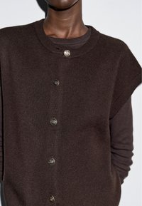 Kobieta nosząca ciemno-brązowy sweter z guzikami, założony na koszulę z długim rękawem, na tle jasnym i jednolitym.