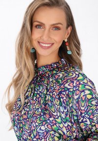 Színes blúz lila háttérrel, élénk zöld, rózsaszín és sárga absztrakt mintázattal, valamint redőzött nyakkivágással.