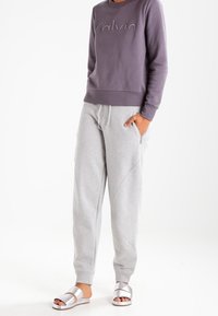 Sudadera gris con el logotipo "Calvin" en relieve, combinada con pantalones de chándal gris claro con costuras diagonales y sandalias metálicas.