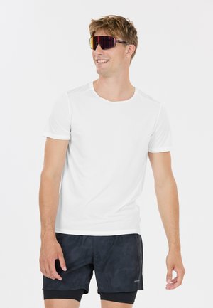 Homme portant un t-shirt blanc, un short de sport noir superposé sur un short de compression noir, et des lunettes de sport réfléchissantes, souriant.