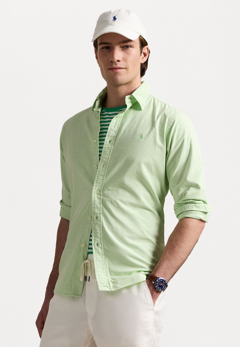 Camisa de botones verde claro superpuesta sobre una camiseta de rayas verdes y blancas, combinada con pantalones de tonos claros y una gorra blanca, mostrando un reloj.