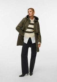 Giacca imbottita verde oliva con zip e bottoni automatici, indossata sopra un maglione a righe crema, abbinata a jeans neri a gamba larga e tacchi neri.