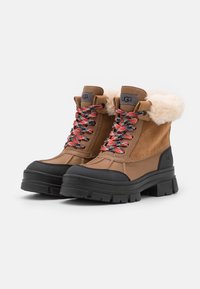 Bottes en daim marron avec un col crème duveteux, une semelle en caoutchouc noire et des lacets à motif rouge et noir. Elles présentent un bout arrondi et une languette rembourrée.