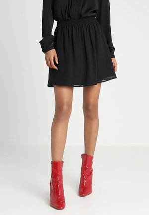 Femme portant une jupe noire et un chemisier à manches longues noir, associée à des bottines rouges brillantes, debout devant un fond blanc uni.