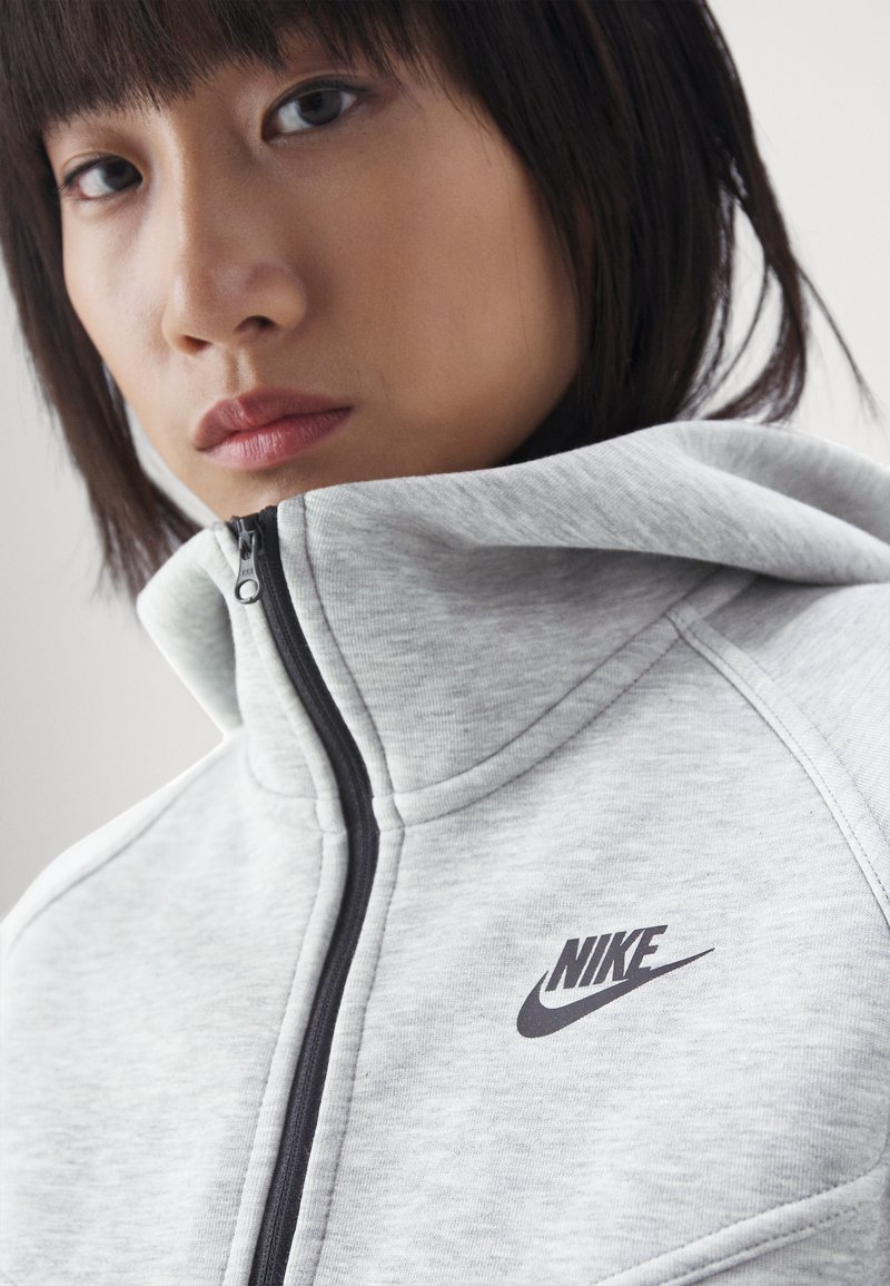 Grauer Zip-Hoodie aus strukturiertem Fleece-Material, mit hohem Kragen, schwarzem Reißverschluss und einem kontrastierenden schwarzen Nike-Logo auf der Brust.