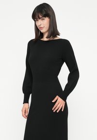 Donna con capelli lunghi e neri lisci, che indossa un abito nero attillato a maniche lunghe, mentre posa con una mano sui fianchi su uno sfondo bianco.