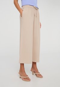 Pantaloni larghi beige chiaro con vita regolabile, tasche laterali, tessuto morbido e una vestibilità rilassata, abbinati a sandali con tacco di colore chiaro.