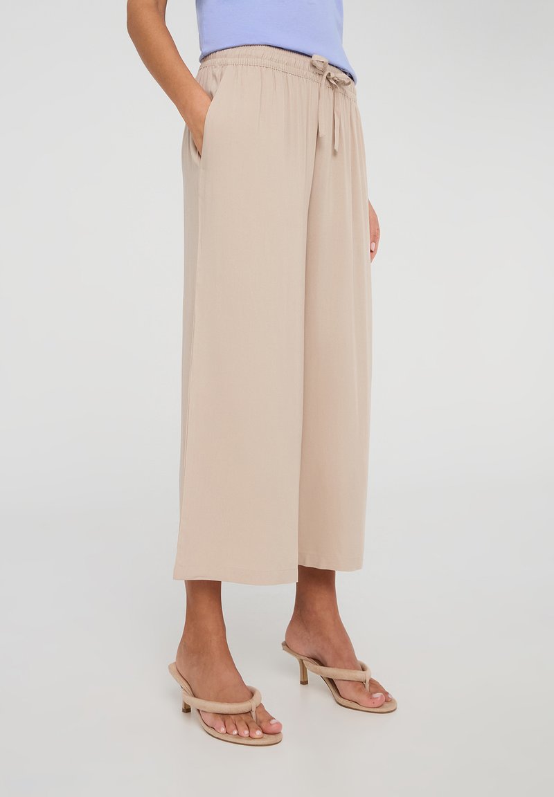 Pantaloni larghi beige chiaro con vita regolabile, tasche laterali, tessuto morbido e una vestibilità rilassata, abbinati a sandali con tacco di colore chiaro.