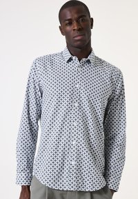 Camisa blanca de botones con un patrón geométrico negro, mangas largas y cuello. La tela parece suave con una textura ligera.