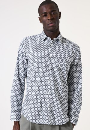 Witte blouse met knopen, voorzien van een geometrisch zwart patroon, lange mouwen en een kraag. De stof lijkt glad met een lichte textuur.