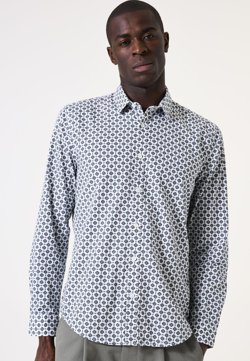 Camisa blanca de botones con un patrón geométrico negro, mangas largas y cuello. La tela parece suave con una textura ligera.