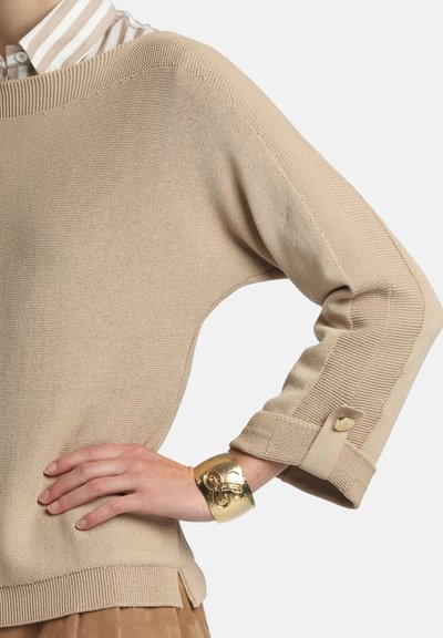 Pull beige en maille avec un col large, manches courtes ornées de boutons, associé à un bracelet manchette texturé doré.