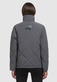 Gray quilted jacket with a high collar and a logo patch on the back. Features a boxy shape and textured surface.