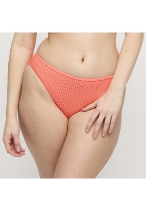 Donna che indossa slip bikini corallo con texture, con la mano appoggiata sulla parte superiore della coscia, su sfondo uniforme.